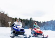 Snøscootersafari To personer kjører hver sin snøscooter på et snødekt underlag, med en bakgrunn av skog og vinterlandskap. Begge har på seg hjelm og kjøreutstyr.