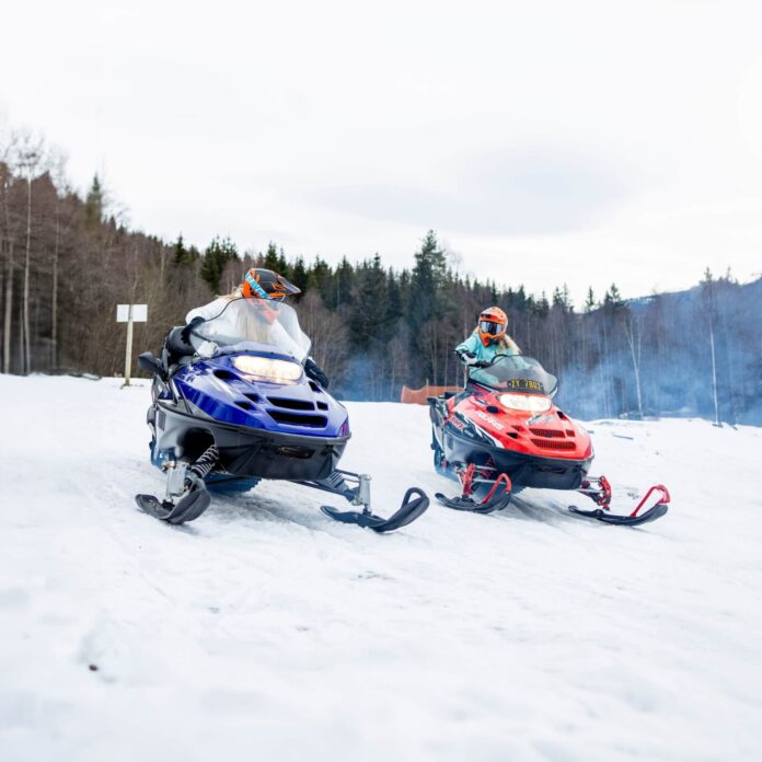 To personer kjører hver sin snøscooter på et snødekt underlag, med en bakgrunn av skog og vinterlandskap. Begge har på seg hjelm og kjøreutstyr.
