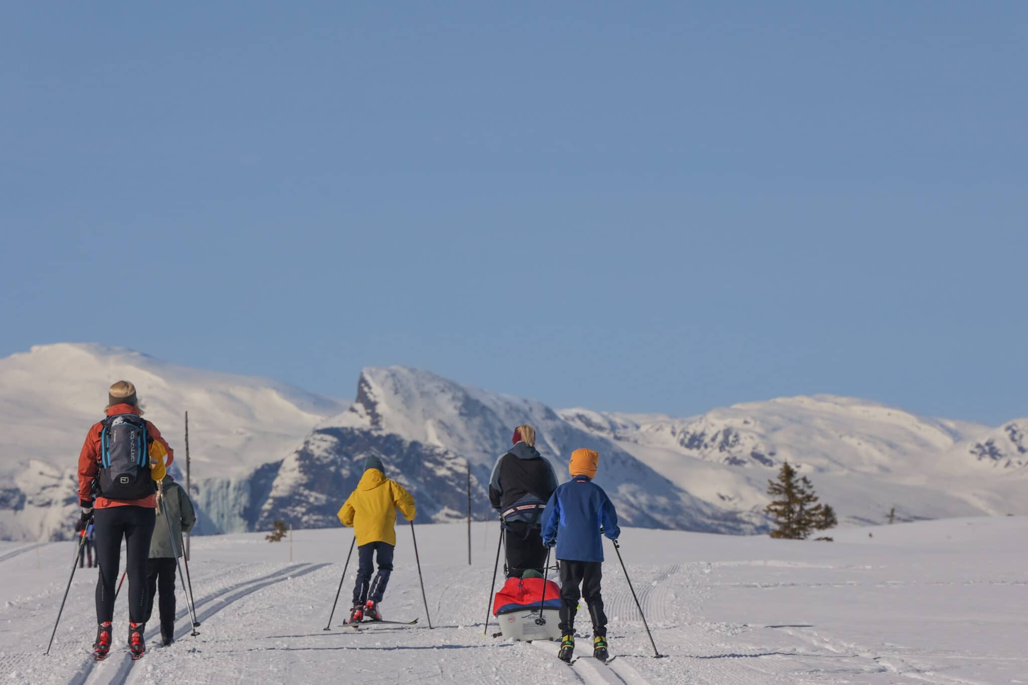 Familie på langrenn i et vinterlandskap med snødekte fjell i bakgrunnen, under en klar, blå himmel. Vinterferie på Oset Fjellhotell Visit Gol